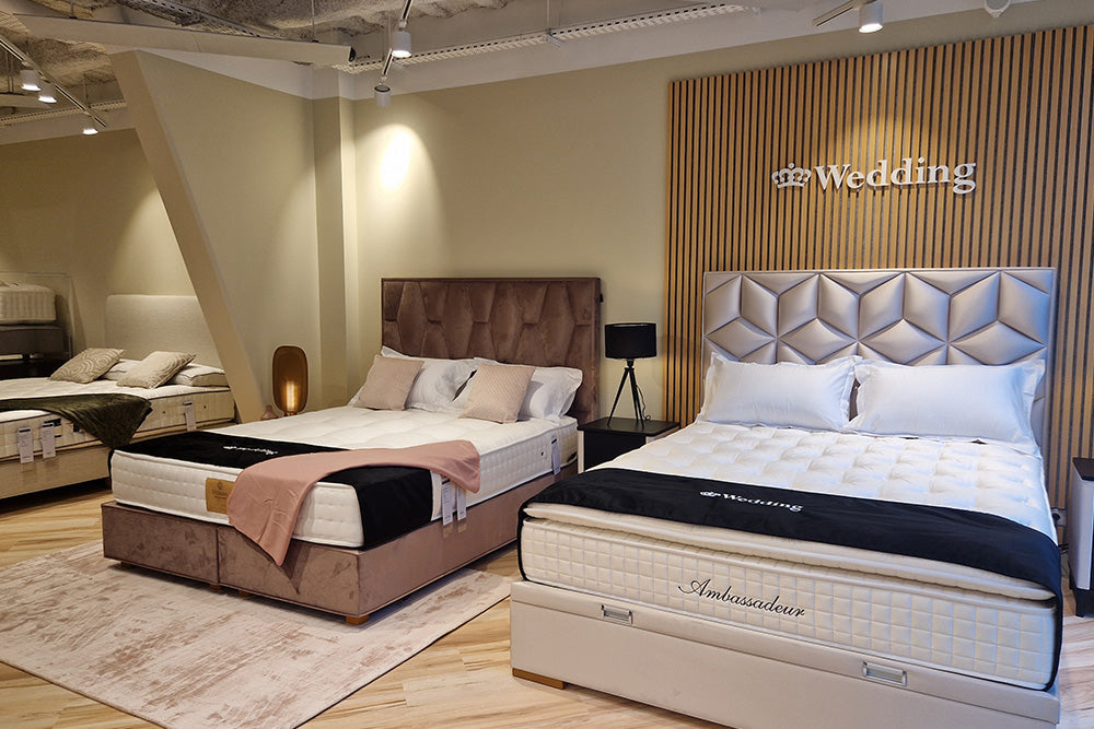 Deux lits élégants exposés dans un showroom, avec des têtes de lit rembourrées et des couvertures marquées "Wedding" et "Ambassadeur"