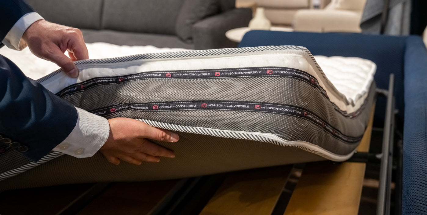 Personne en costume examinant un matelas dans un magasin, avec l'étiquette "La Maison du Convertible" visible