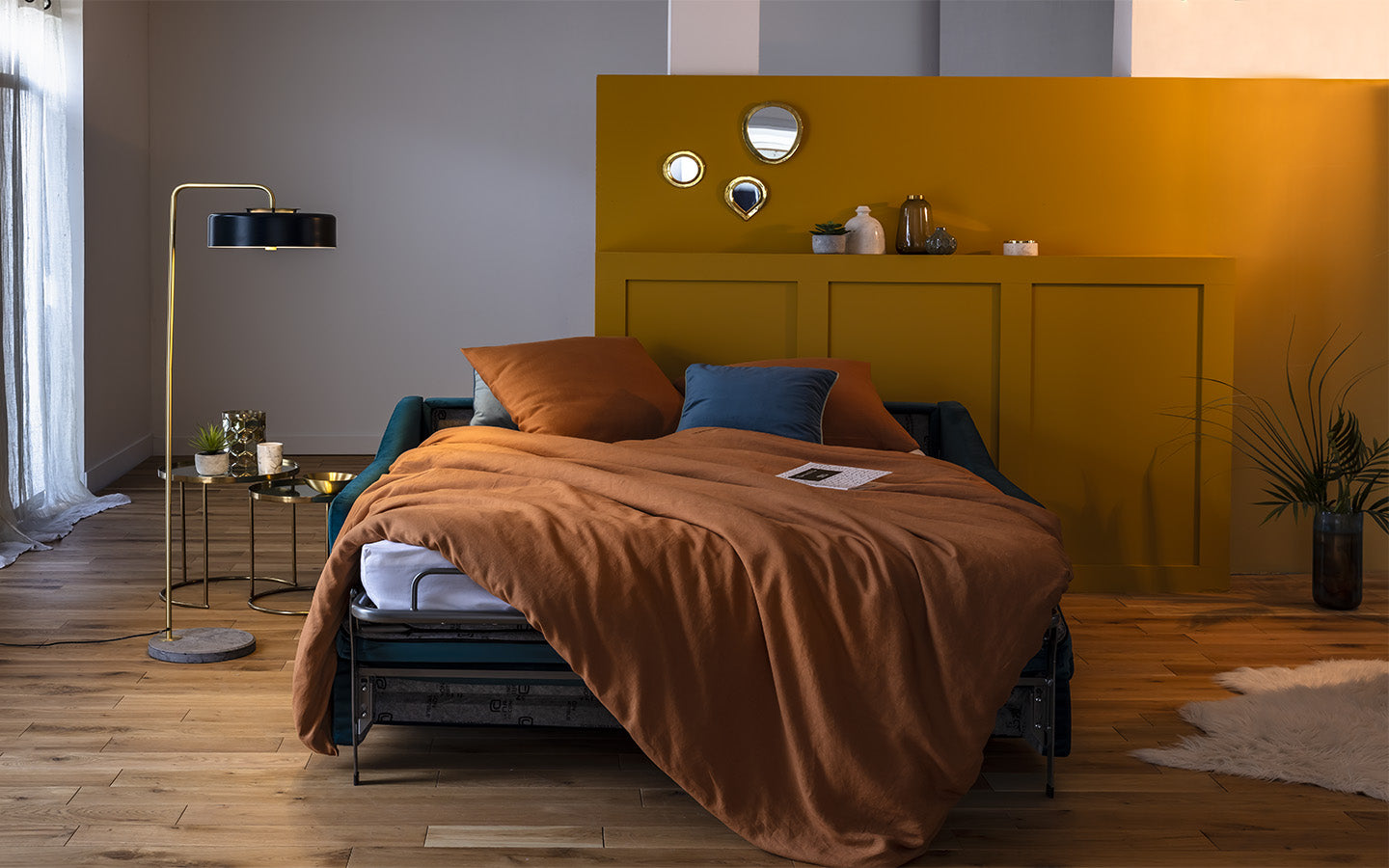 Canapé convertible Layne déplié en lit, ambiance nuit avec couverture orange, lampadaire doré et mur jaune en arrière-plan