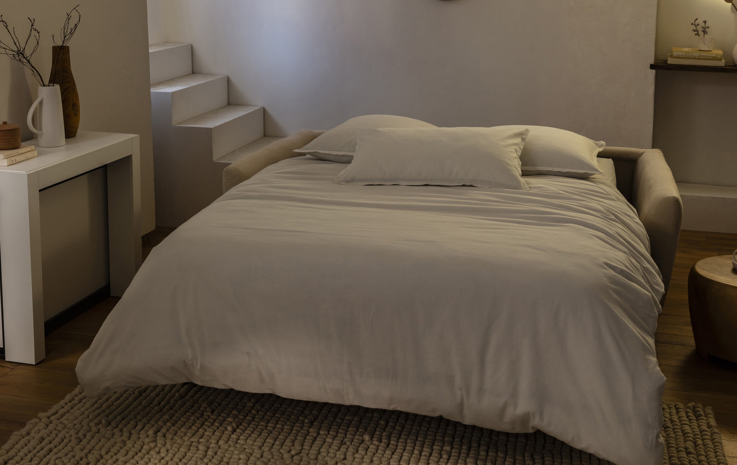 Canapé convertible beige en tissu, ambiance nuit, avec coussins, sur un tapis texturé dans un intérieur moderne et épuré