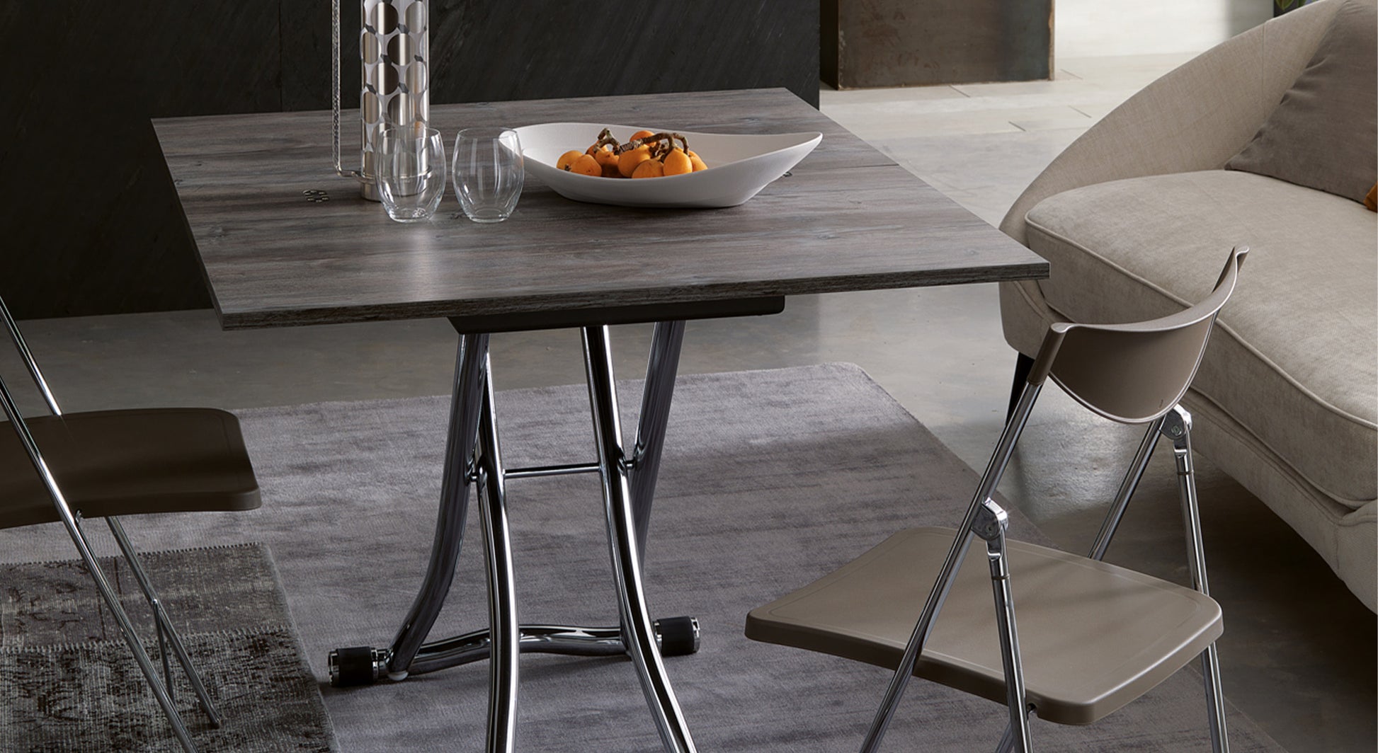 Table extensible moderne ouverte avec plateau en bois, entourée de chaises pliantes et d'un canapé, décorée d'un plat de fruits